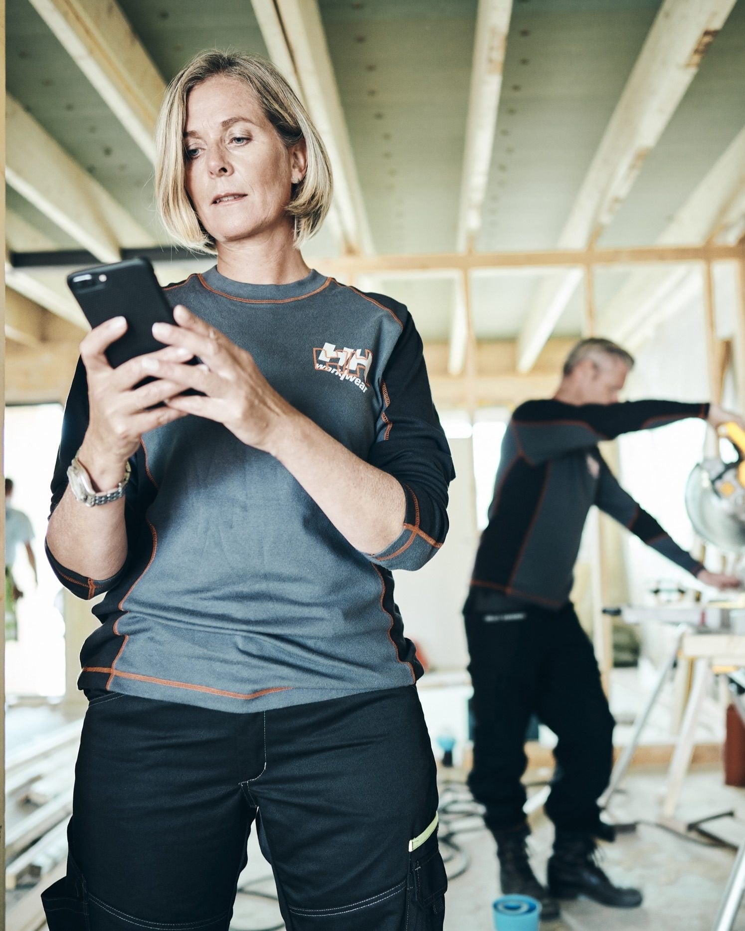 A carpenter checks her phone in a timber-frame building while a colleague cuts at a mitre saw behind her.