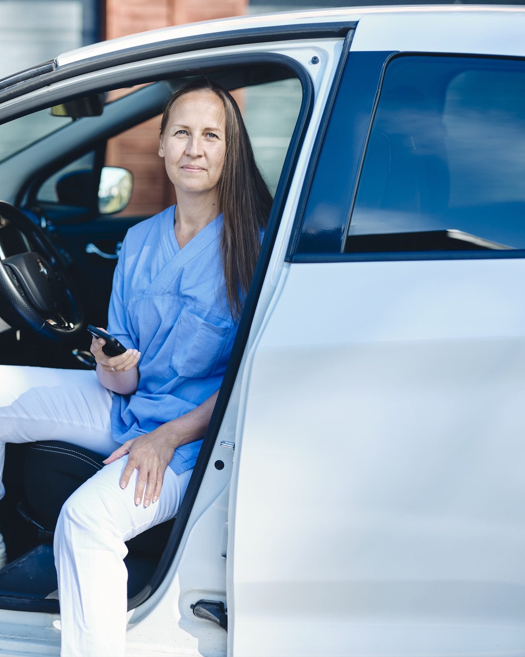 A woman in a blue work tunic sits in the open door of a white car, phone in hand, between jobs.
