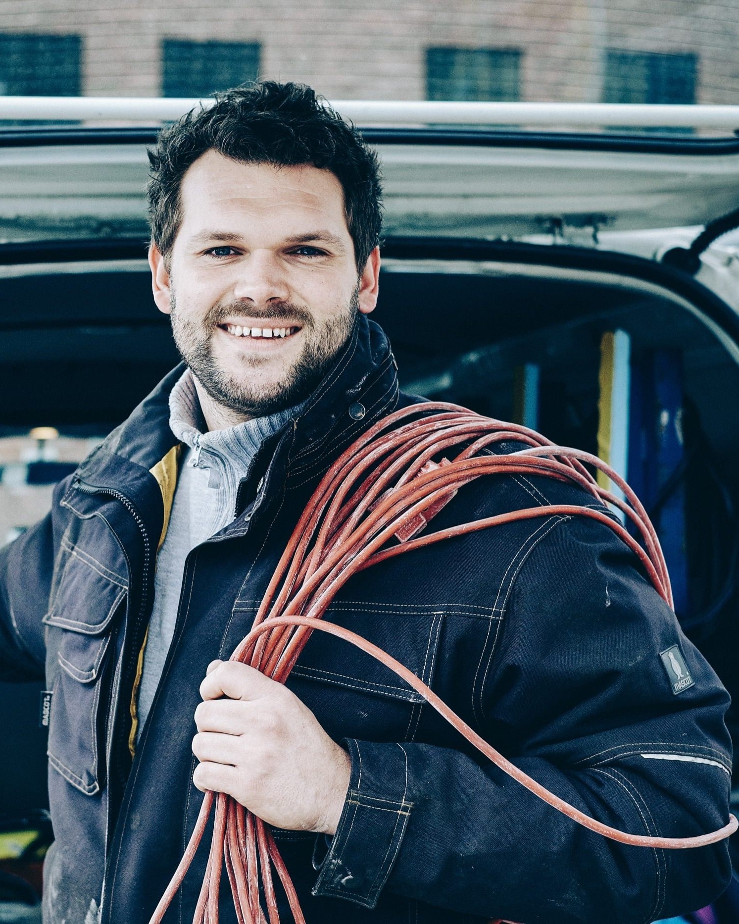 An electrician with a coil of cable over his shoulder at the back of his work van.
