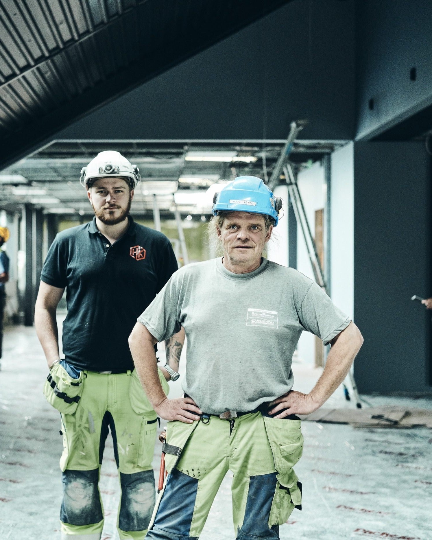 Two painters in work trousers stand in a commercial space under renovation, with colleagues working behind them.