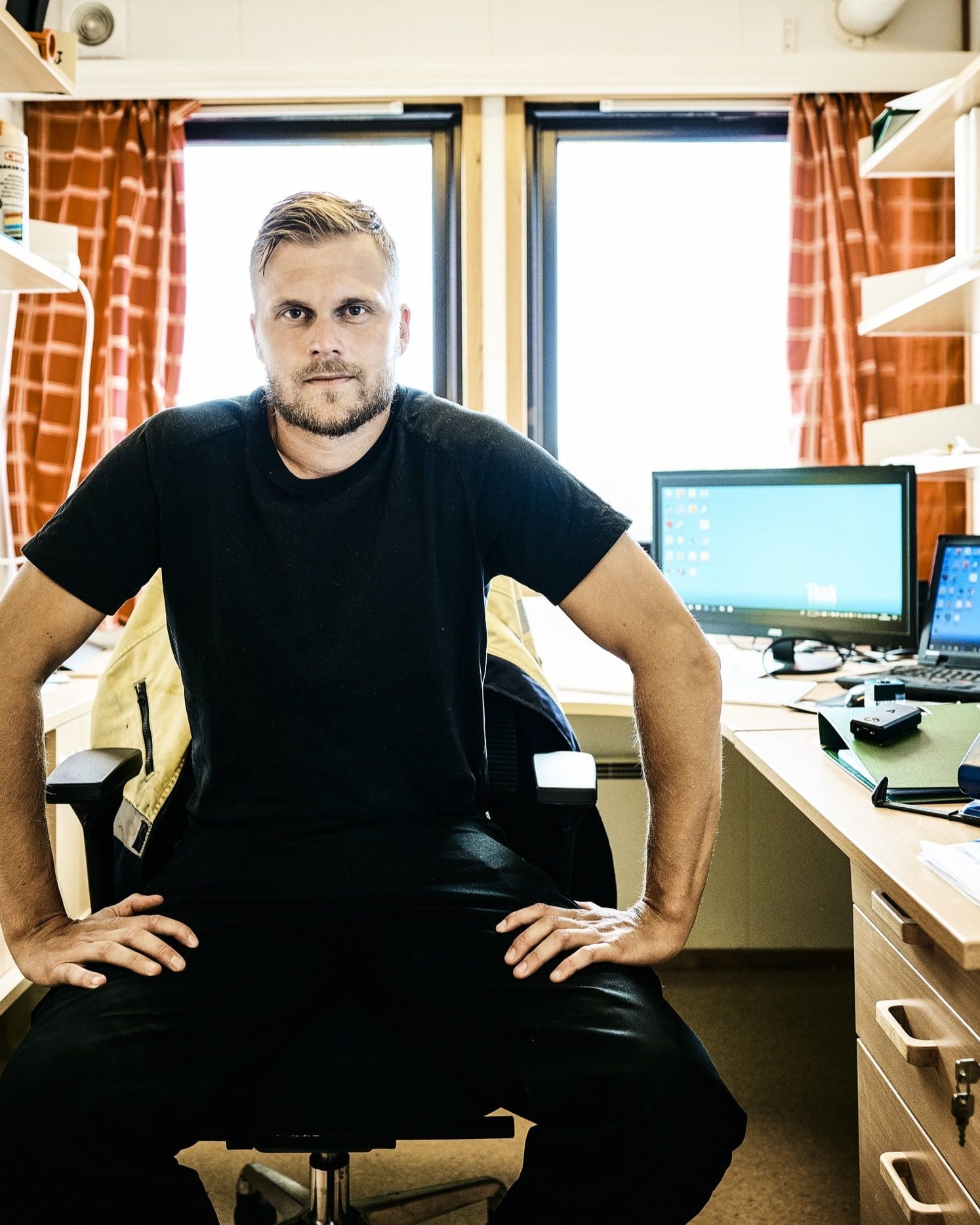 A building operations manager sits at his desk in a site office, with monitors, binders and a hard hat around him.