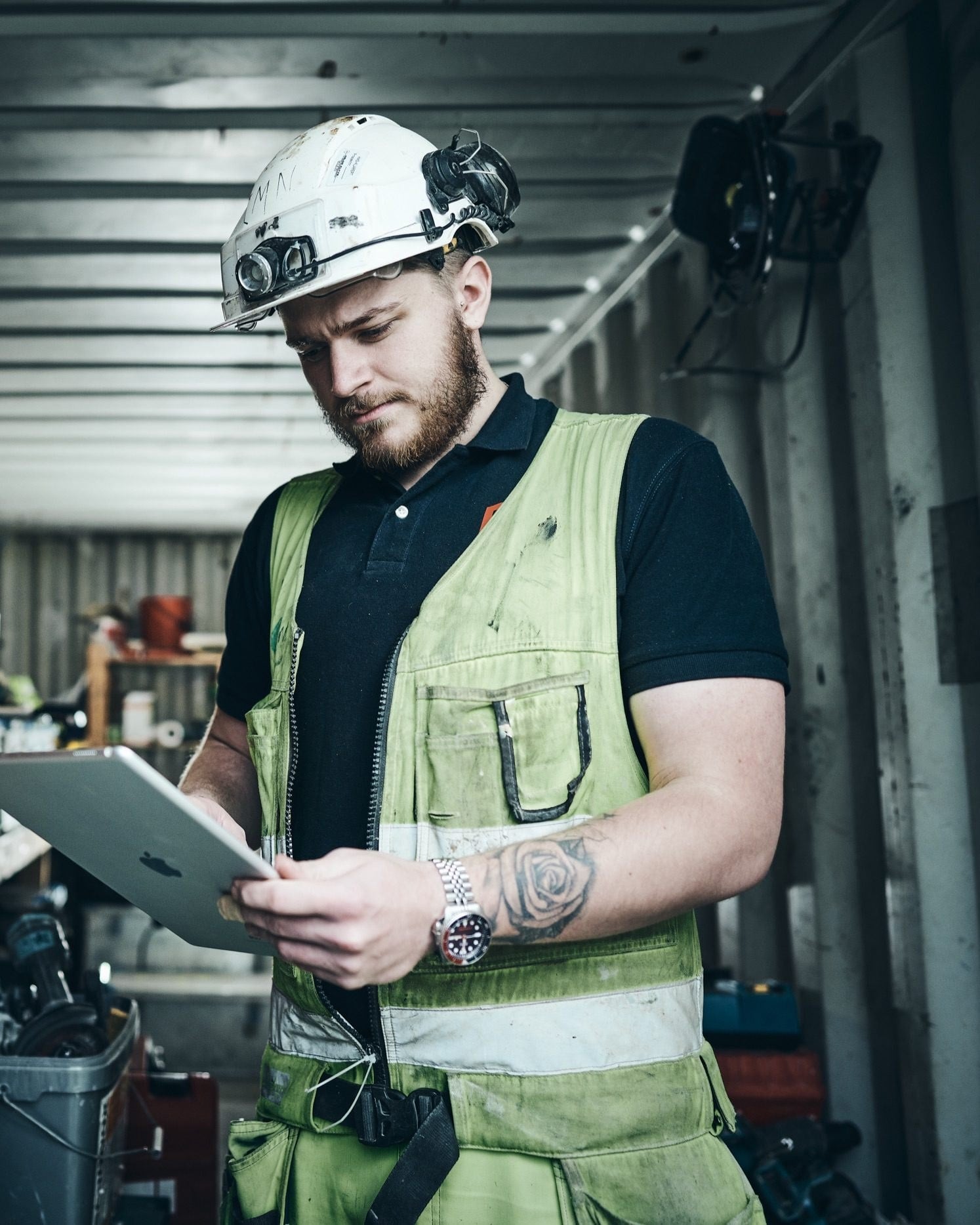An HVAC technician in a hard hat and hi-vis vest reads a job on a tablet in a site container.